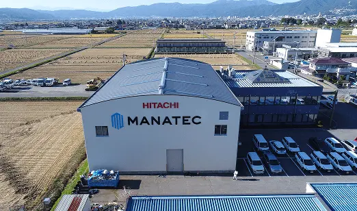 田畑に囲まれた場所に建つ工場とオフィスの外観。建物にはHITACHI MANATECのロゴが表示されている。